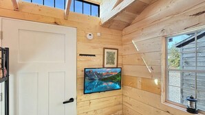 A wooden cabin interior with a white door, a mounted TV, a heater, and a window with a view of the outdoors. - Zen Box Farm (Bomoseen)