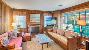 A cozy living room with wooden paneling, a fireplace, a sofa set, a coffee table, and a large window with a view. - Raven's Nest (Ogunquit)