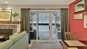 A living room with a fireplace, a sofa, a dining table, and a view of a snowy deck through glass doors. - Gateway Condo A10 (Rutland)