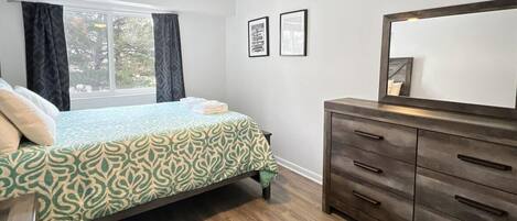 A bedroom with a bed, a dresser, a ceiling fan, a window with curtains, and framed pictures on the wall.