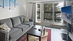 A living room with a grey sofa, a coffee table, a flat-screen TV, and a large window with a view of trees and a deck. - Mt Green 2-d07 (Killington)