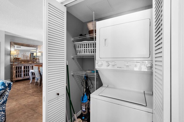 A laundry room with a front-loading washer and dryer, shelves with baskets, and a mirror above a cabinet. - Portofino Island Resort 1-1402 (Pensacola Beach)