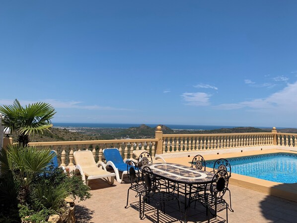 Enjoy a serene coastal view from the sunlit patio, featuring a pool, table, and chairs, surrounded by a low wall and balustrade.
