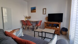 A living room with a sofa, a dining table with chairs, a TV on a wooden stand, and a window with blinds. - Beautiful renovated apartment in the heart of Concarneau - Private parking - WIFI (Concarneau)