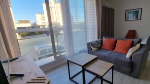 A modern living room with a grey sofa, a small wooden table, and a view of the city through large windows. - Beautiful renovated apartment in the heart of Concarneau - Private parking - WIFI (Concarneau)
