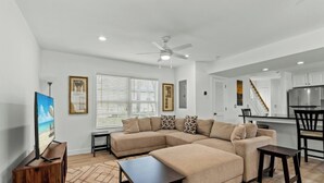 A modern living room with a sectional sofa, a coffee table, and a TV. There is a kitchen area with a fridge and a staircase. - Sand At The Strand-Pool, Cabana, 1/2 Mile To Shops (North Myrtle Beach)