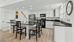 A modern kitchen with white cabinetry, a stainless steel refrigerator, a microwave, and a large island with chairs. - Sand At The Strand-Pool, Cabana, 1/2 Mile To Shops (North Myrtle Beach)