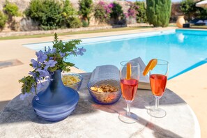 Een zwembadterras met een bloevas, schalen met snacks en twee drankjes op een tafel.