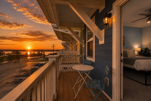 A balcony with a view of the sunset over a body of water, a small table, and a bedroom with a bed and nightstand.