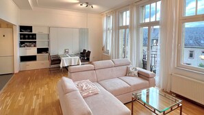 Un espacio habitable amplio con un sofá en forma de L, una mesa de centro de vidrio y una mesa de comedor con sillas. Hay armarios empotrados y vistas al exterior a través de grandes ventanales.