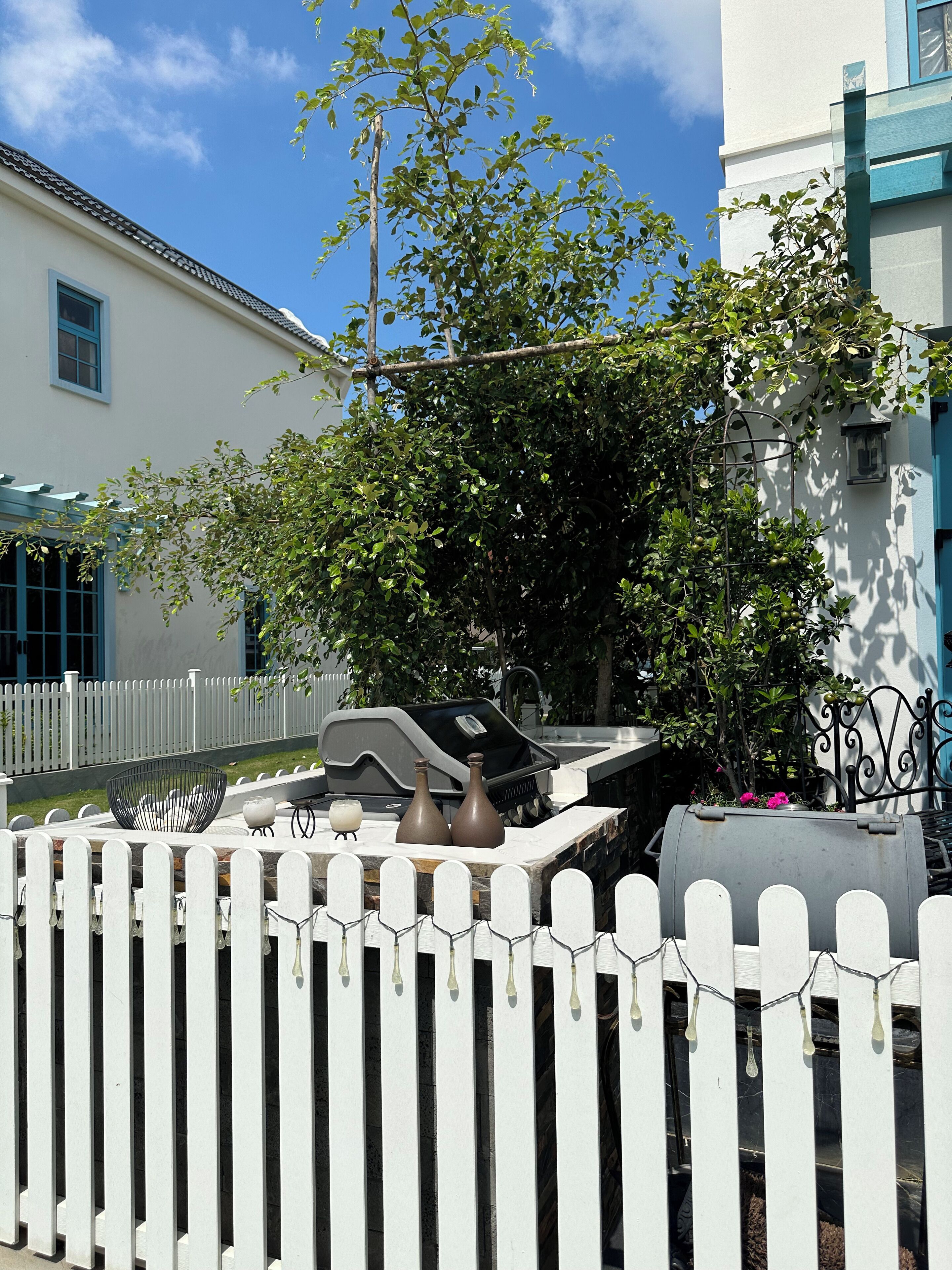A barbecue grill with a white picket fence in the foreground, a tree, and a building with a blue door.