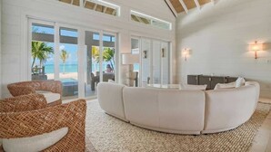 Een ruime zithoek met een grote witte bank, rieten stoelen en uitzicht op het strand door grote schuifdeuren.