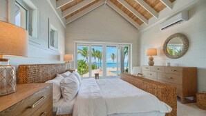 A bedroom with a large bed, wooden headboard, a dresser, a vanity with a mirror, and a view of the beach through a sliding glass door.