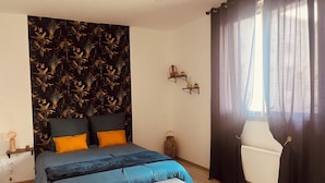 Une chambre à coucher comprenant un lit, une table de chevet, une lampe, une fenêtre avec des rideaux et un mur recouvert d’un papier peint à motifs tropicaux.