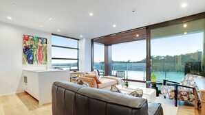 A modern living room with a large sofa, a dining area, and a view of a body of water through large windows. - Gorgeous Home with Catalina Bay Views and Carpark (Auckland West)