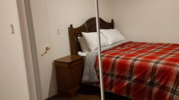 Een slaapkamer met een houten hoofdbord, een rode geruite spreidekker, een houten nachtkastje en een wit kussen.