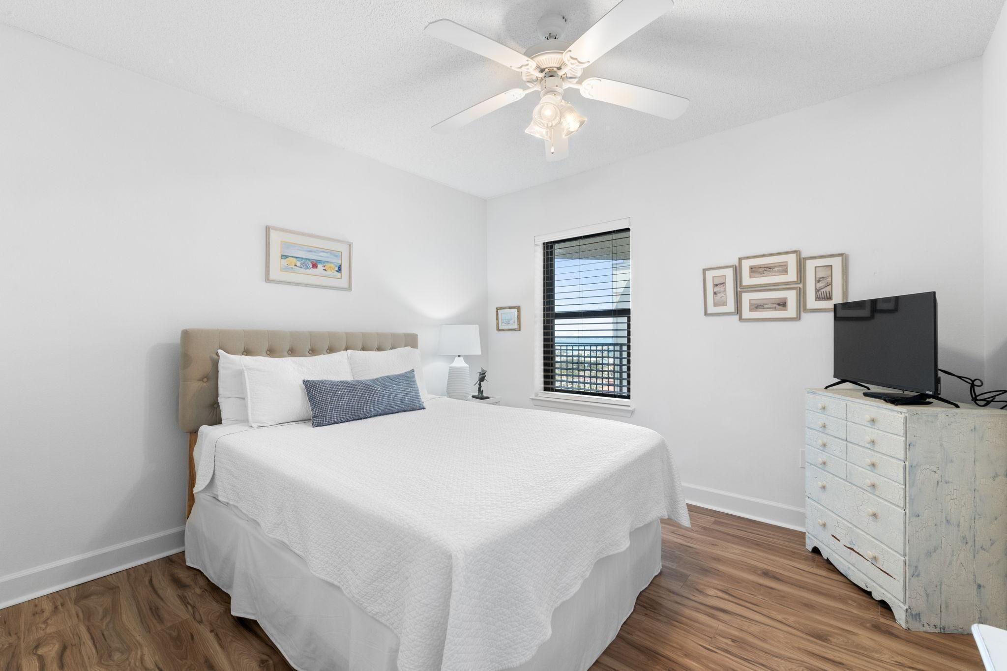 Une chambre à coucher avec un lit, un ventilateur de plafond, une commode et une fenêtre avec des stores.