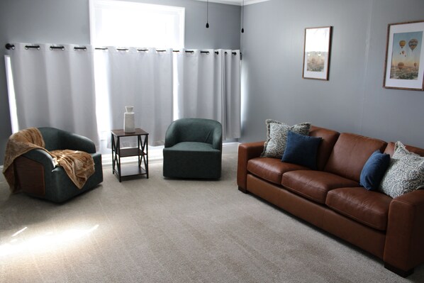 Un salon avec un canapé en cuir marron, un fauteuil vert et un fauteuil foncé. On y trouve une petite table sur laquelle sont posés une lampe et un objet blanc. Les murs de la pièce sont de couleur gris clair et il y a une grande fenêtre avec des rideaux blancs.