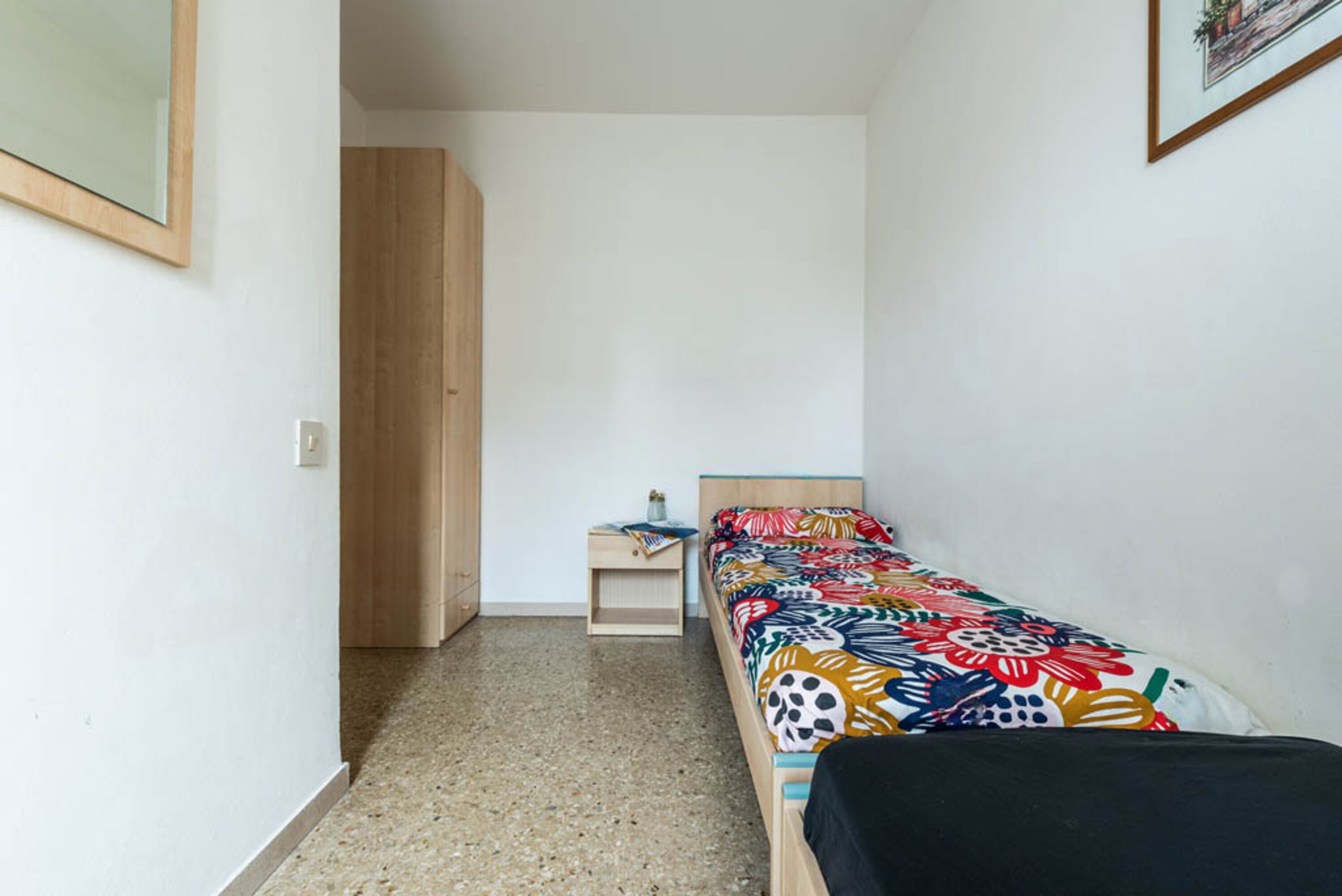 A small, narrow room with a single bed, a nightstand, and a wooden wardrobe.