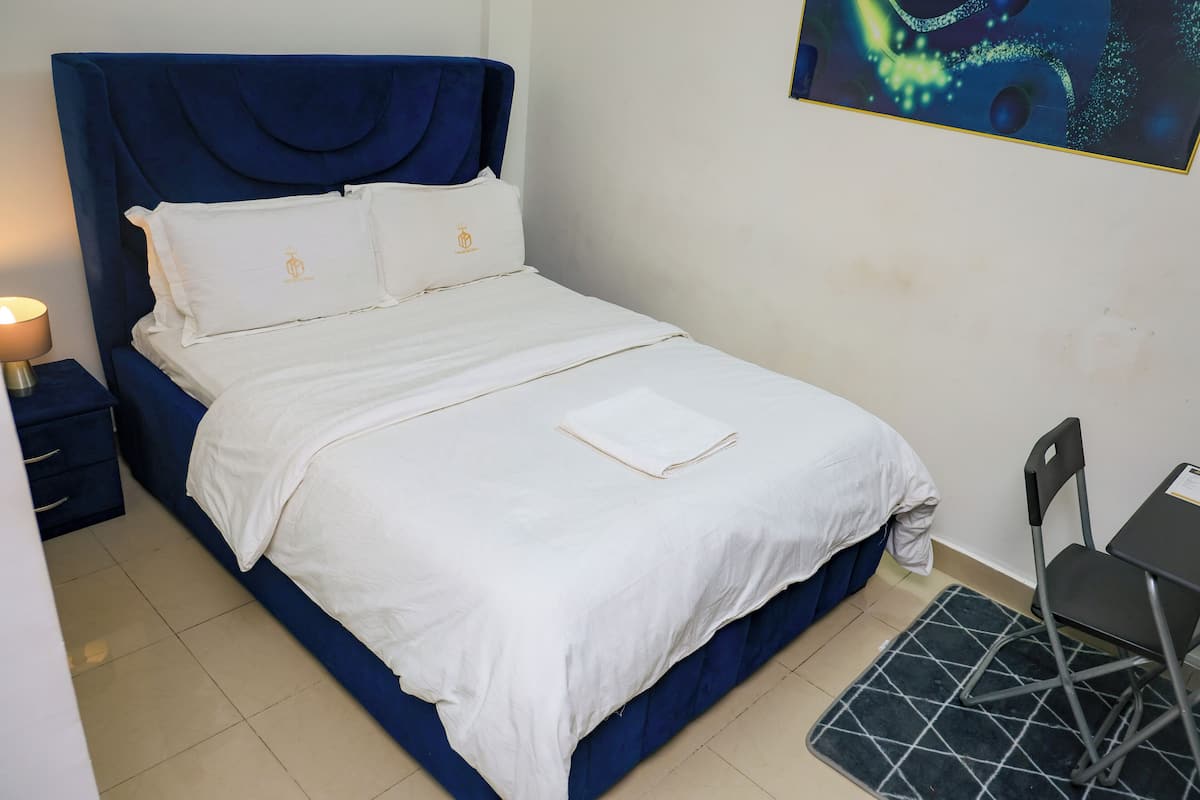 A neatly made bed with white linens and a blue headboard, a small bedside table with a lamp, and a foldable chair.