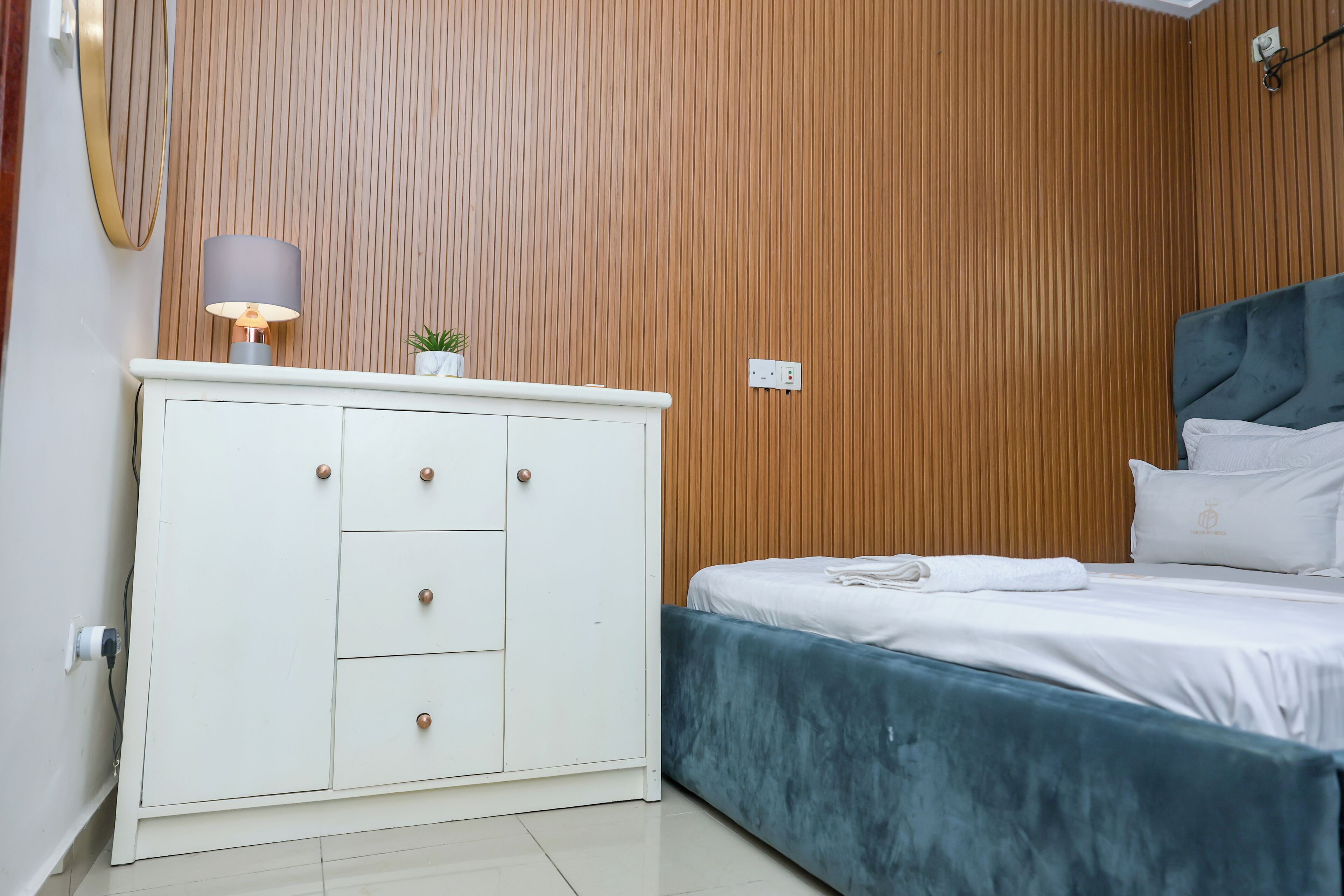 A bedroom with a wooden panel wall, a white dresser with drawers, a bed with a blue headboard, and a bedside lamp.