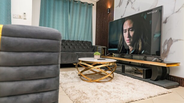 A modern living room with a grey sofa, a round coffee table with a gold base, a flat-screen TV on a wooden stand, and a white fluffy rug. - T-Square Residence Makepe (Douala)