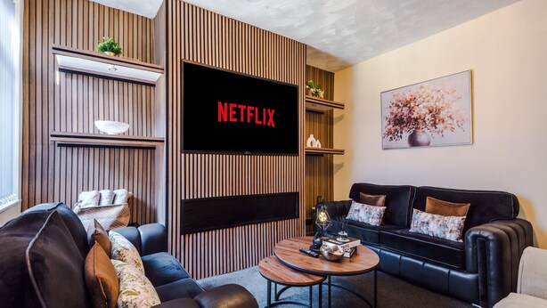 Ein modernes Wohnzimmer mit einem Flachbildfernseher, der 'NETFLIX' anzeigt, einer schwarzen Ledersofa, einem Holztisch und einer dekorativen Vase.
