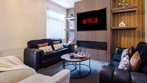 Ein modernes Wohnzimmer mit einem flachbildfernseher an einer Holzwand, einem schwarzen Ledersofa, einem runden Couchtisch und einem Bücherregal mit Dekoration.