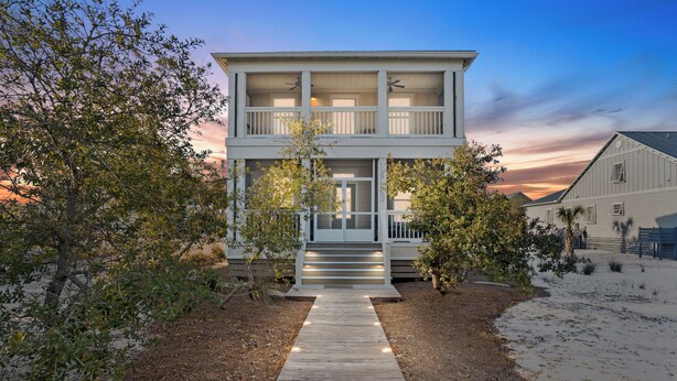 A two-story house with a wooden deck leading to the front door, surrounded by trees and a sandy area. - 1st Tier 3BR Home w/ Pools, Golf Cart & Beach (Port St. Joe)