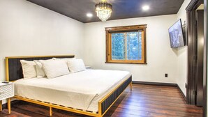 A bedroom with a large bed, a chandelier, a window with a view of trees, and a television on the wall.