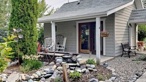 A small, single-story house with a porch, a stone pathway, and a garden with a variety of plants and a tree. - Cottage Suite at Country Way Inn (Kalispell)