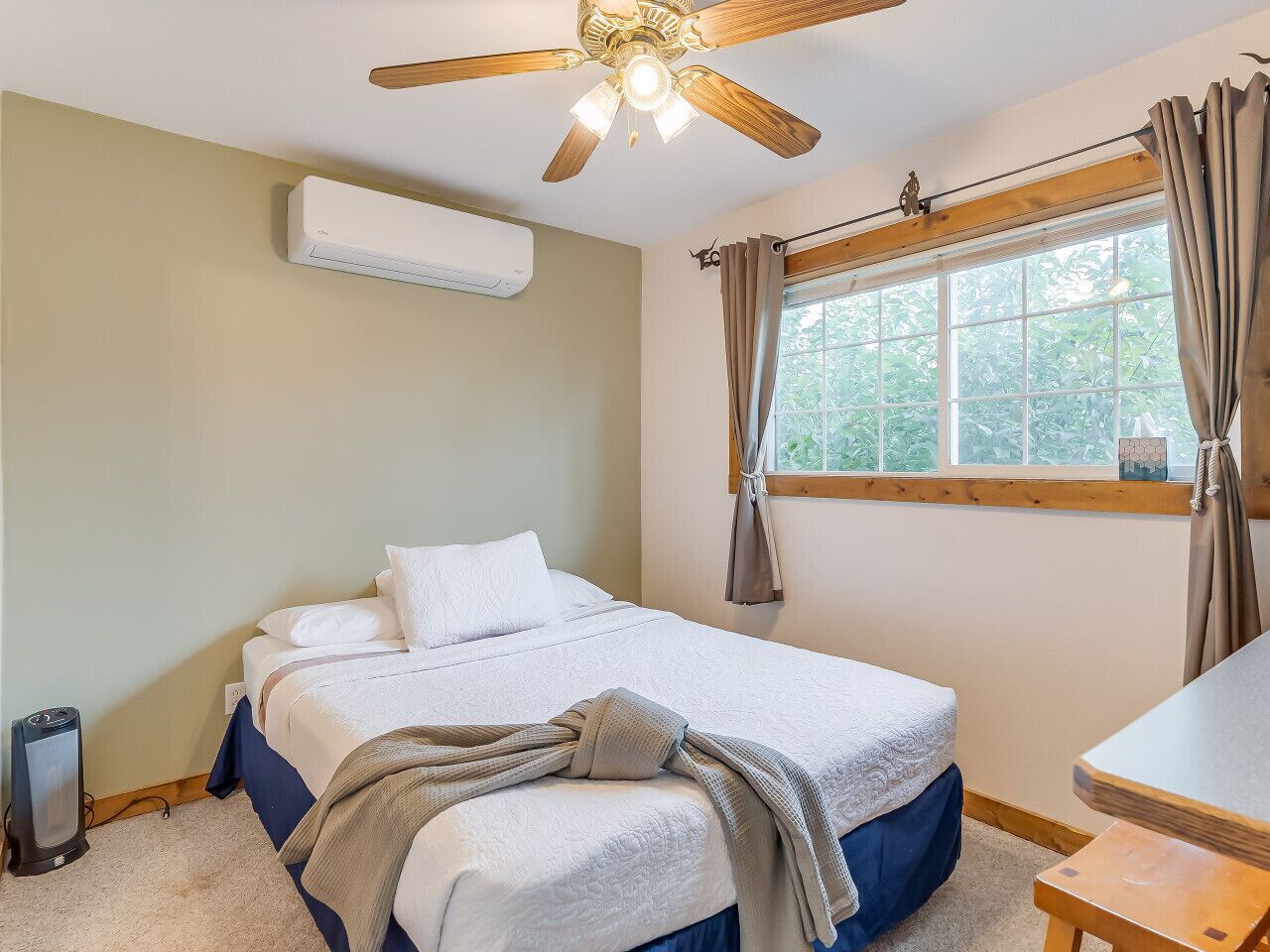 A bedroom with a bed, a ceiling fan, an air conditioning unit, a window with curtains, and a bedside table.