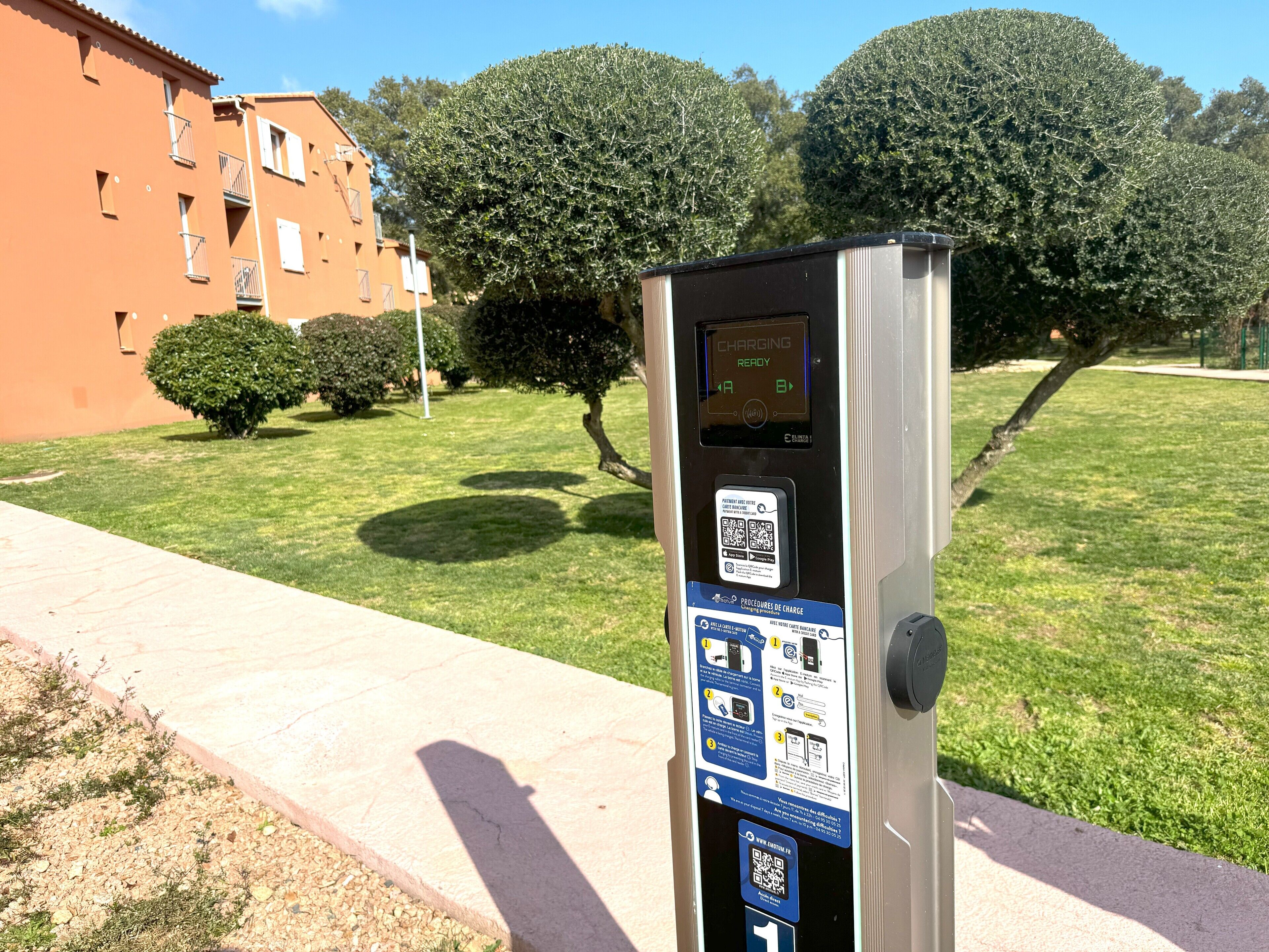 Une borne de recharge publique, munie d’instructions et d’un code QR, située sur un trottoir, avec un bâtiment et des arbres en arrière-plan.