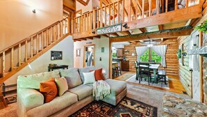 Ein Blockhaus-Interieur mit einer Holztreppe, einem Essbereich und einem Wohnzimmer mit einer Couch und einem gemusterten Teppich.