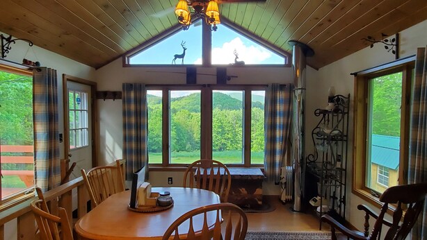 A dining area with a wooden table and chairs, a ceiling fan, and large windows offering a view of a forested landscape. - "Pleasant Brook Cottage" near Roxbury Village - Woods, Streams, Wi-fi, sleeps 6 (Roxbury)