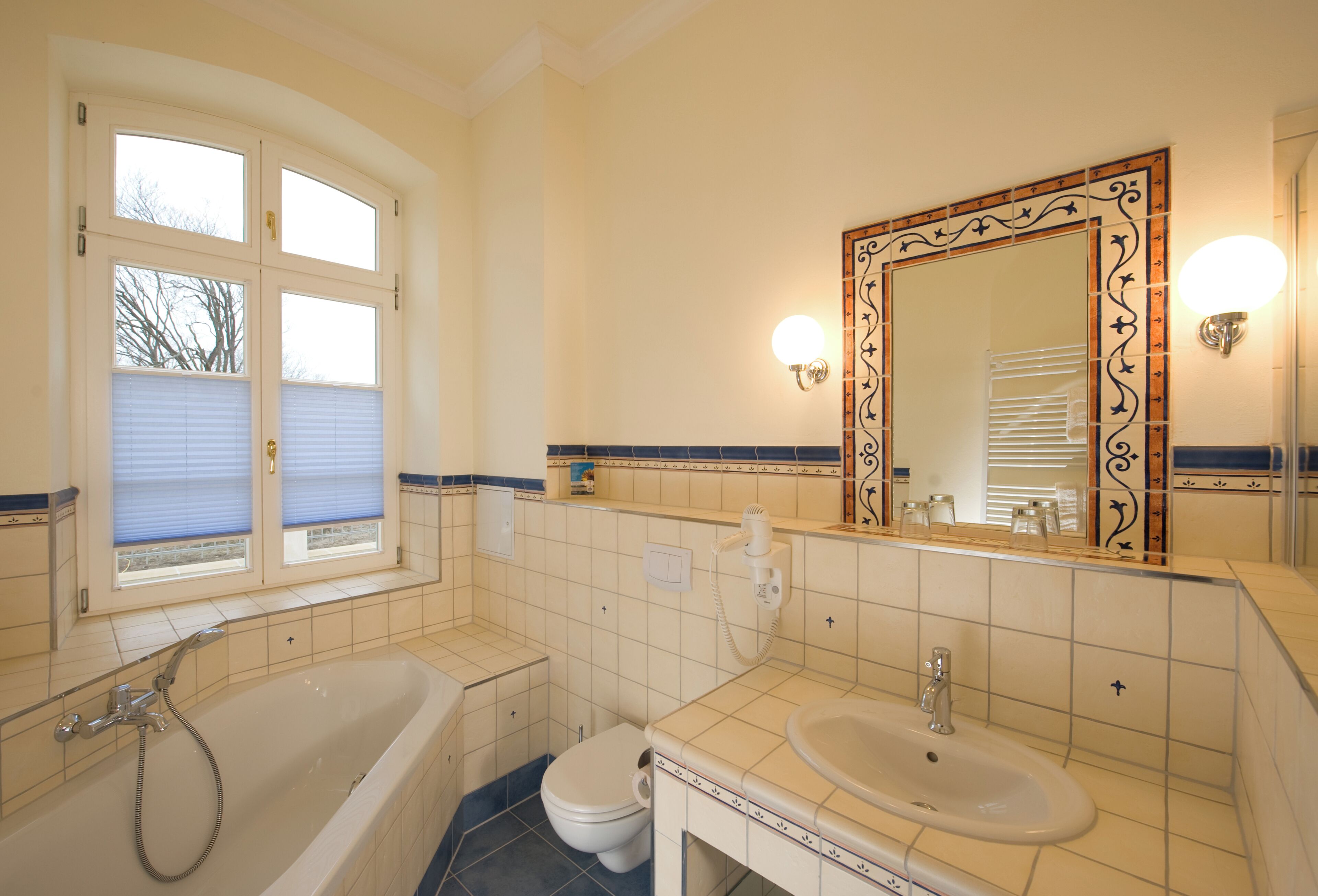 Un baño con bañera, inodoro y lavamanos, que incluye un espejo grande con detalles decorativos e iluminación en la pared.