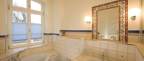 Un baño con bañera, inodoro y lavamanos, que incluye un espejo grande con detalles decorativos e iluminación en la pared.