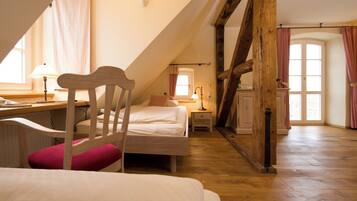 Ein Schlafzimmer mit schrägen Wänden, Holzbalken, einem Bett, einem Stuhl, einem Schreibtisch und einem Fenster mit Vorhängen.