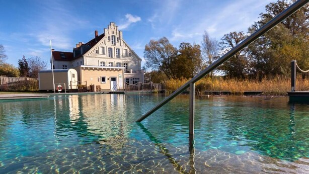 A clear pool with a metal railing, a large building with a gabled roof, and trees in the background. - Bio- und Gesundheitshotel Gutshaus Parin (Damshagen)