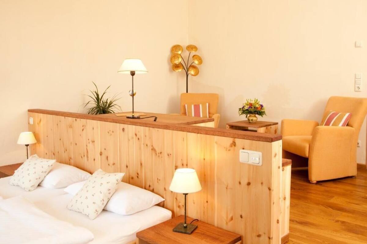 Ein hölzernes Bett mit weißer Bettwäsche und zwei Nachttischlampen, ein Holzerrezeptionstresen mit einer Lampe, ein Sessel mit Kissen und ein dekoratives Wandbild.