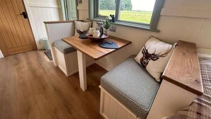A cozy room with a wooden table, two chairs, a bench with a patterned cushion, and a window with a view of a green field. - Luxury Shepherds Hut set in a Countryside Setting (Southampton)