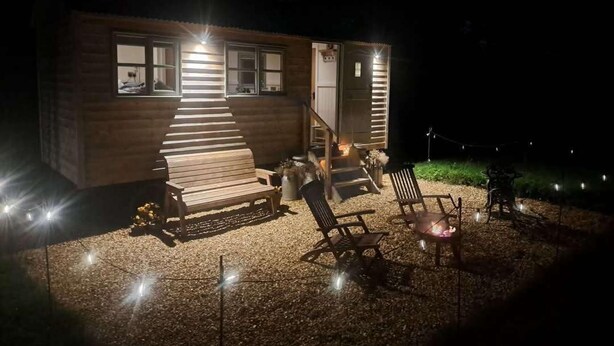 A wooden cabin with a staircase leading to a deck, outdoor seating, and a fire pit. - Luxury Shepherds Hut set in a Countryside Setting (Southampton)