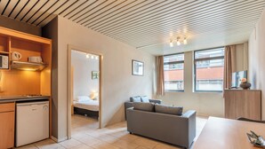 A modern kitchen with a built-in microwave, a small refrigerator, and a wooden cabinet. There is a gray sofa and a wooden dining table in the living area. The room has large windows with curtains and a view of a brick building outside. - Room-one Bedroom Apartment Near North S (Brussels)