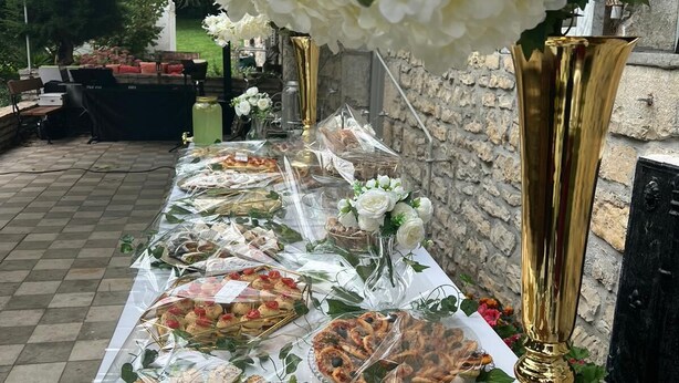 A buffet table with various dishes wrapped in plastic, surrounded by white flowers and gold vases.