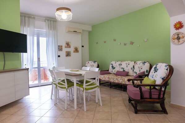 A dining area with a table and chairs, a sofa with floral upholstery, and a television mounted on the wall.