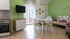 Una cocina moderna con horno empotrado, comedor con mesa y sillas blancas, sofá con tapicería floral y un televisor montado en la pared.