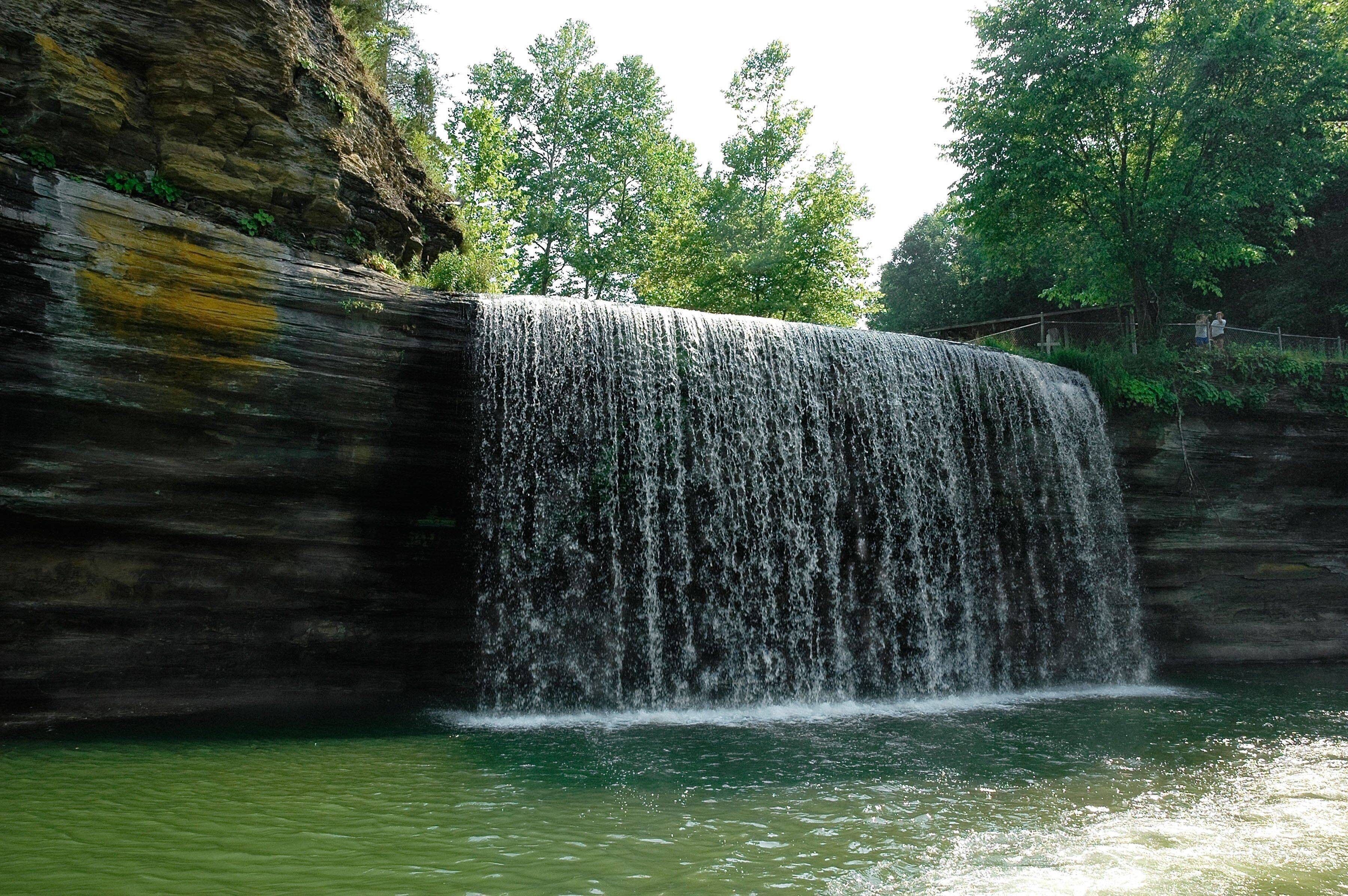 Een waterval die over een rotsachtige klif stroomt in een waterpartij, omgeven door weelderige begroeiing.