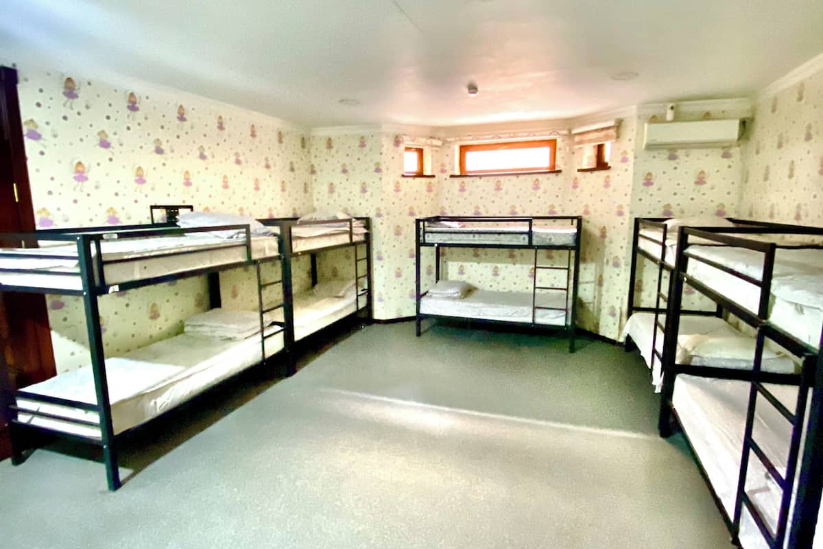 A dormitory room with bunk beds arranged in a U-shape, a window with a wooden frame, and a ceiling light fixture.