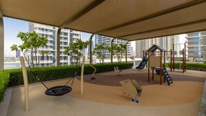 An outdoor play area with swings, slides, and climbing structures, surrounded by a covered seating area and greenery. - Waterfront Living w Unparalleled Sea Palm Vws (Dubai)