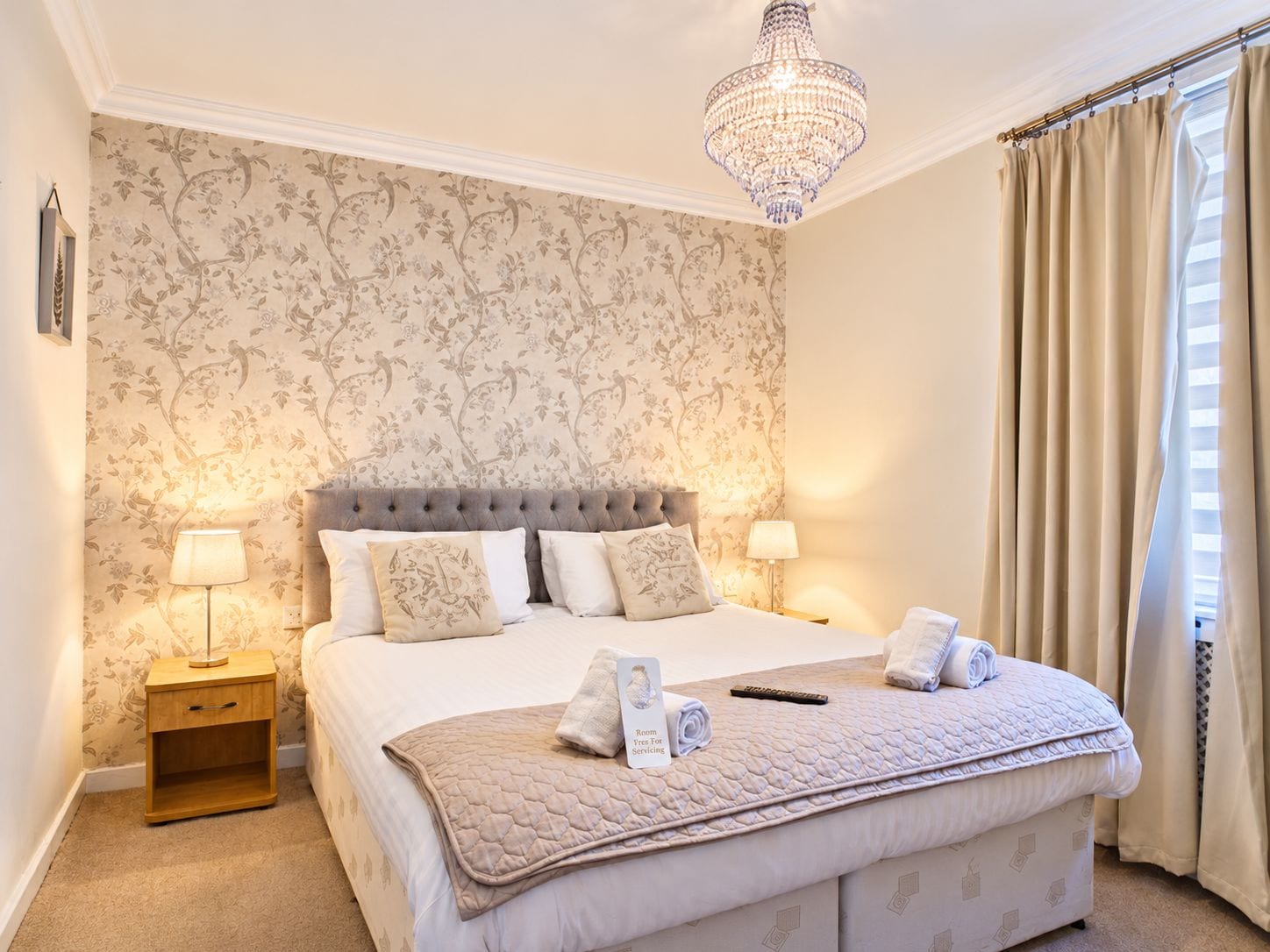 A hotel room with a large bed, two bedside lamps, a chandelier, and a window with curtains.